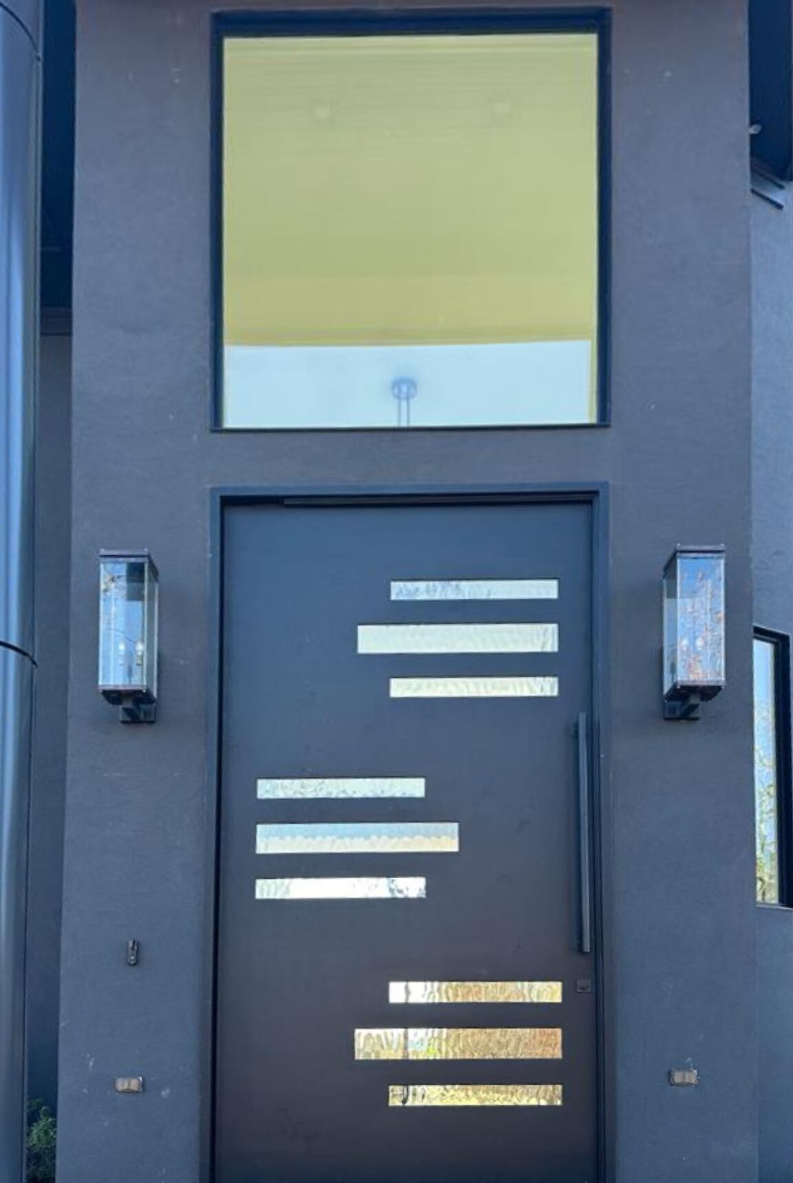 Modern front door with a matte black finish featuring horizontal frosted glass inserts of varying lengths, flanked by two rectangular wall-mounted glass light fixtures.